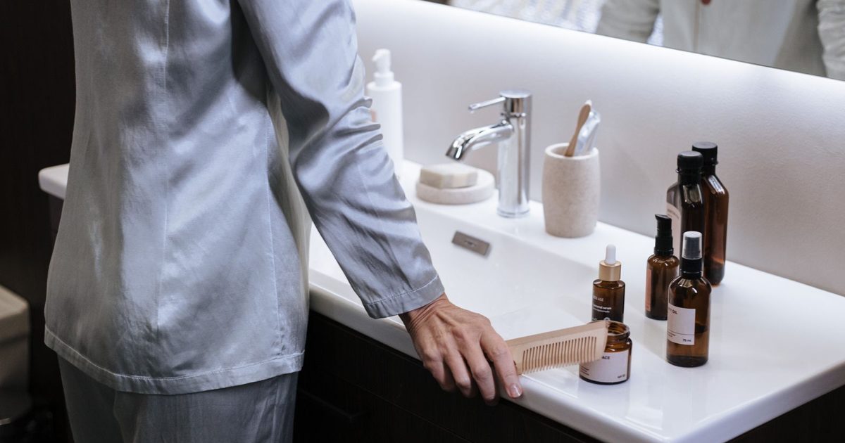 An older woman in satin pyjamas choosing skincare products at a bathroom sink.