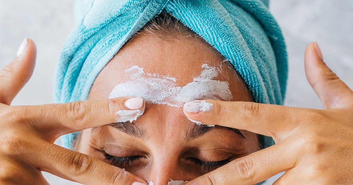 A woman with a turquoise towel applying facial cream to her forehead.