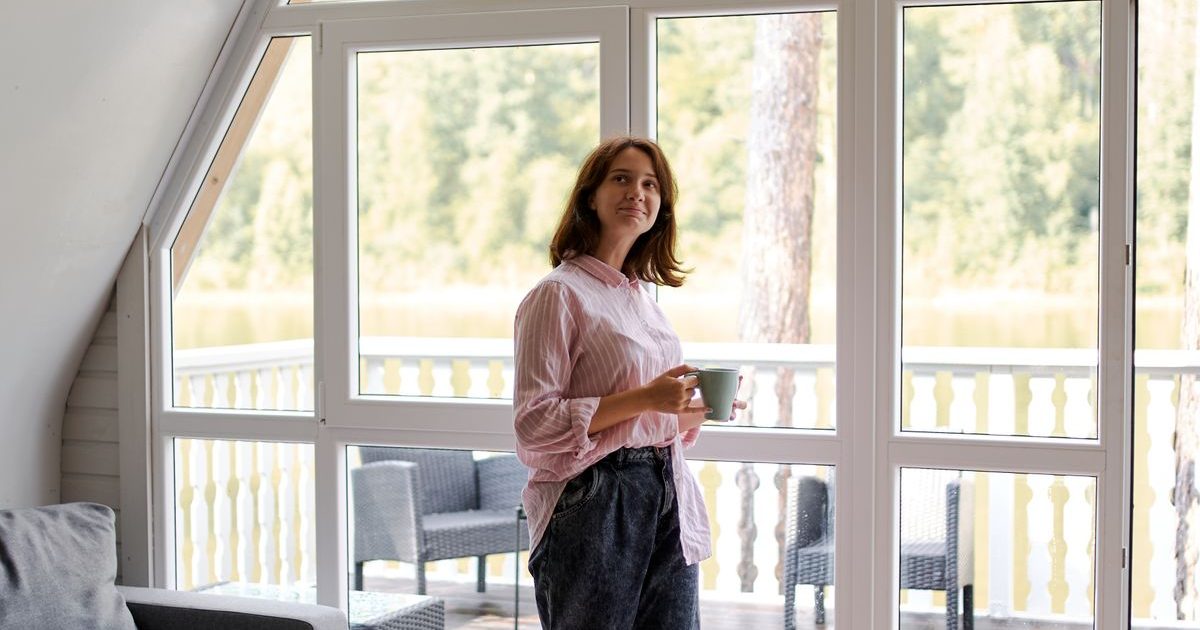 A woman standing by a large window holding a cup of tea.